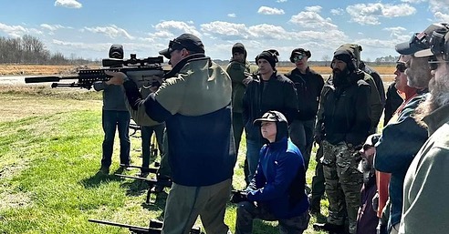 Group of civilians watching a suppressed rifle demonstration at an outdoor range
